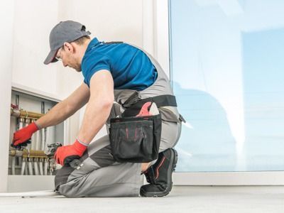 Ein Techniker mit Kappe, blauem Hemd und Arbeitsoverall kniet, um Sanitärrohre in einer Wandverkleidung zu installieren.