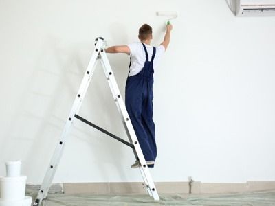 Eine Person in blauem Overall steht auf einer Leiter und streicht eine weisse Wand mit einer Farbrolle.
