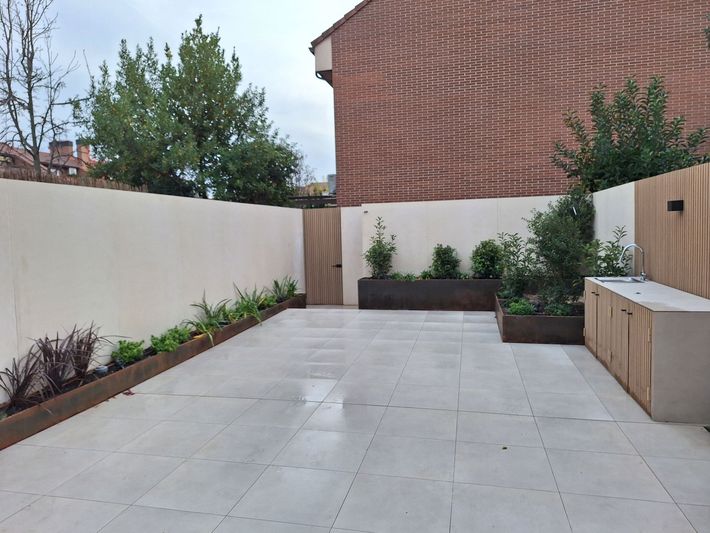 Un patio moderno con suelo de baldosas claras y una cocina exterior integrada. Jardineras con vegetación adornan las paredes.