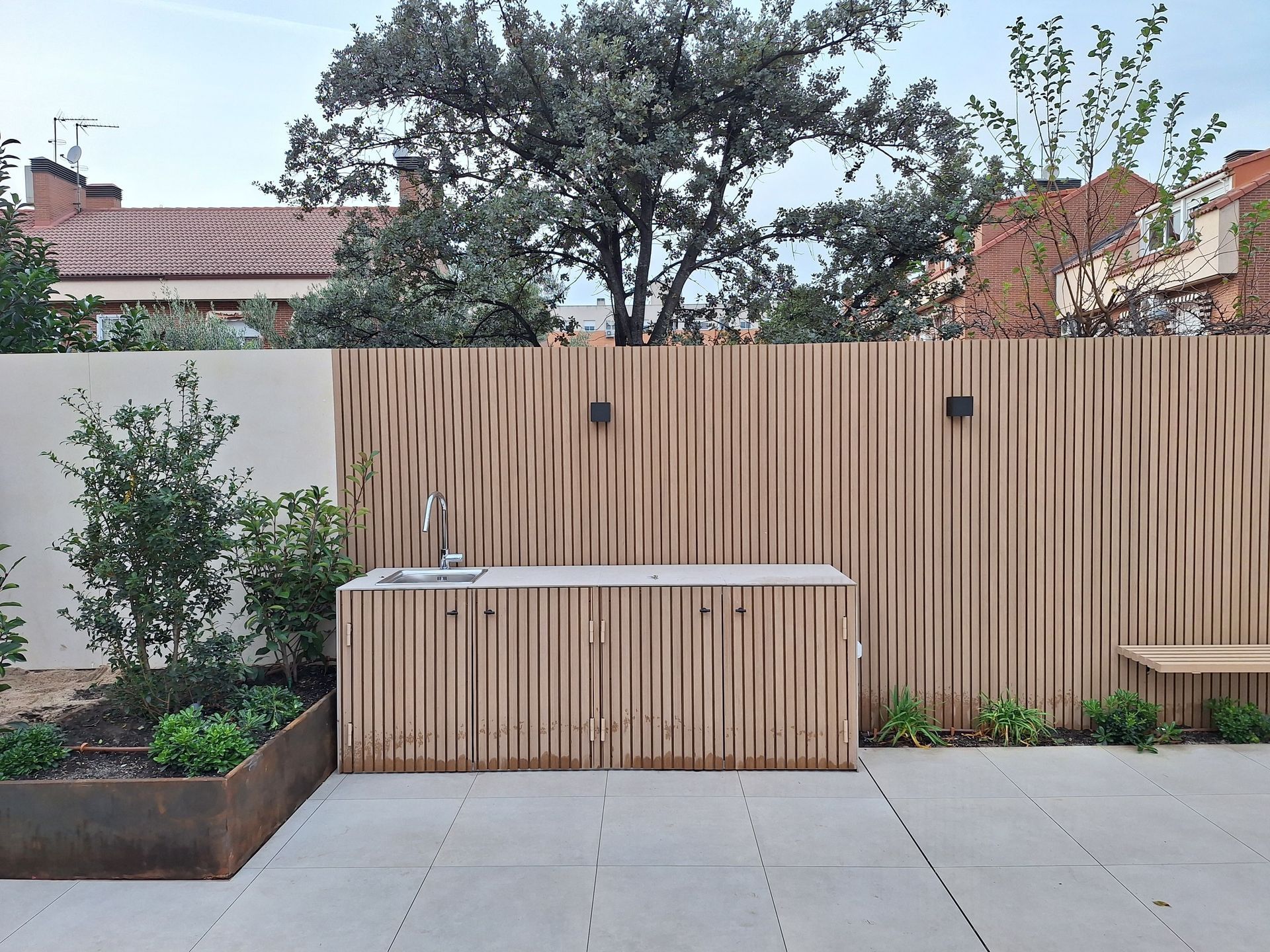 Cocina exterior con valla de madera y encimera. Patio de baldosas grises con vegetación y árboles al fondo.