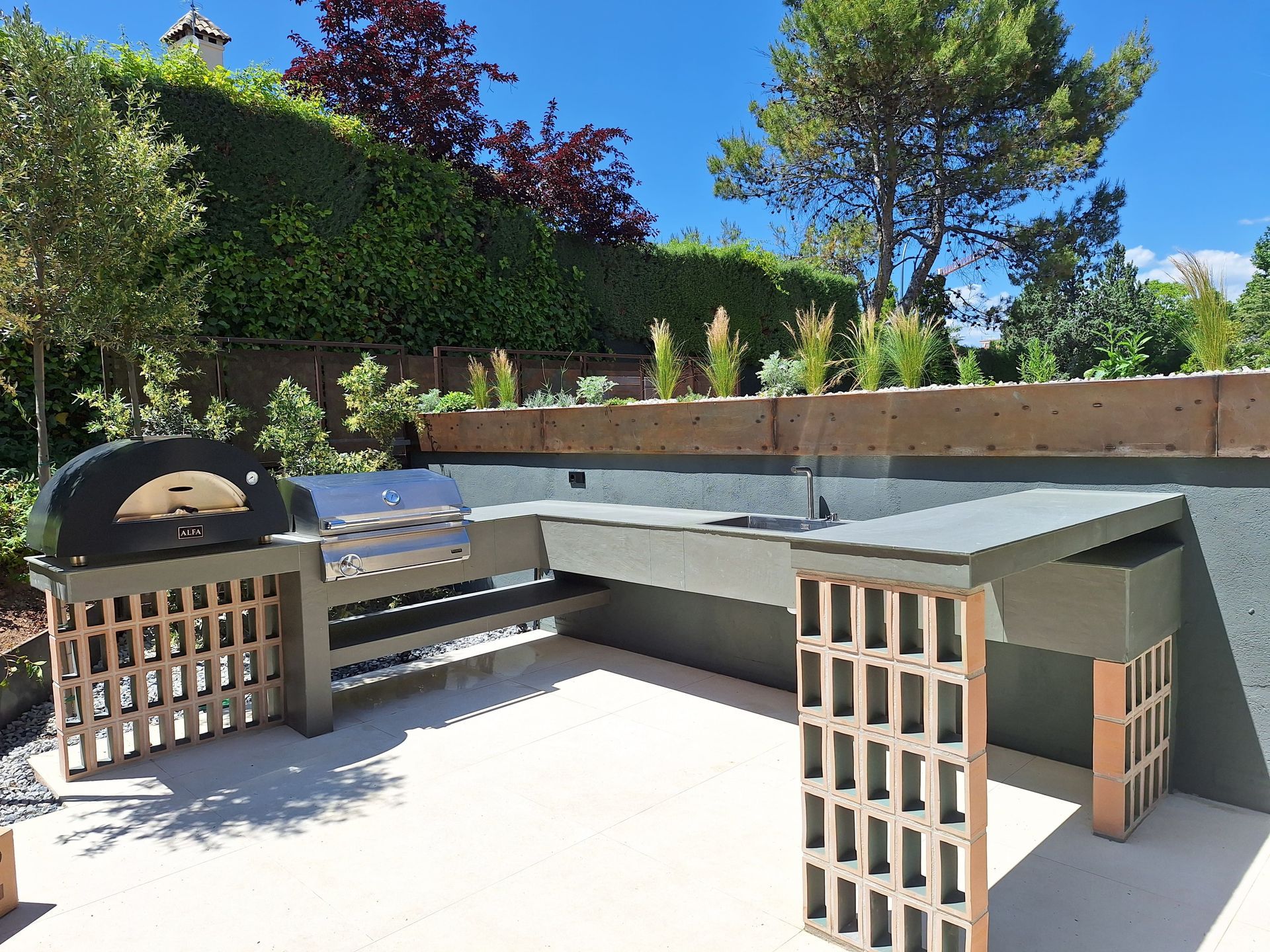 Cocina al aire libre con horno de pizza, parrilla y encimera en un patio, rodeada de vegetación y un muro de piedra.