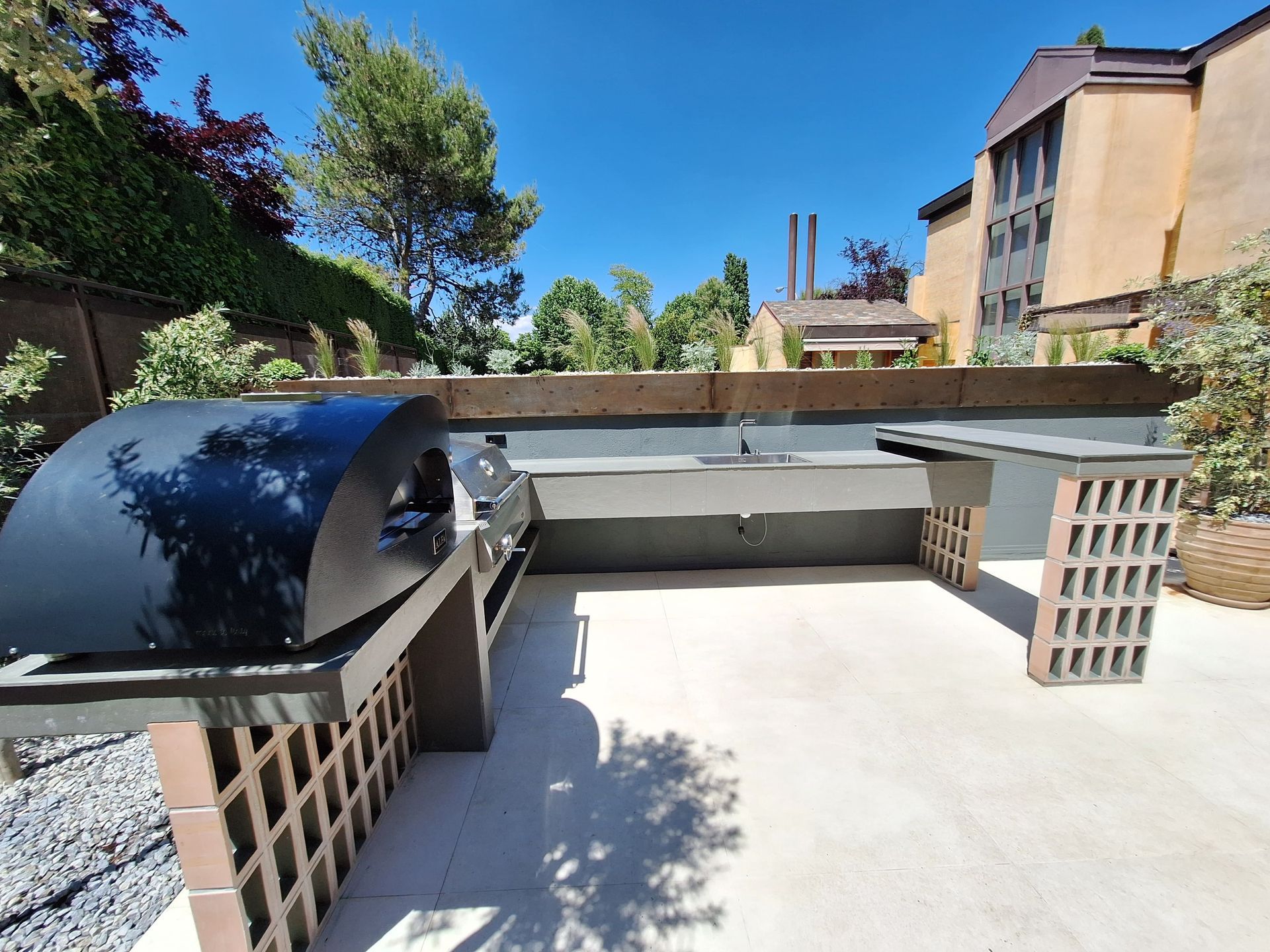 Cocina al aire libre con un horno de pizza negro y encimeras grises contra un fondo soleado con un edificio.