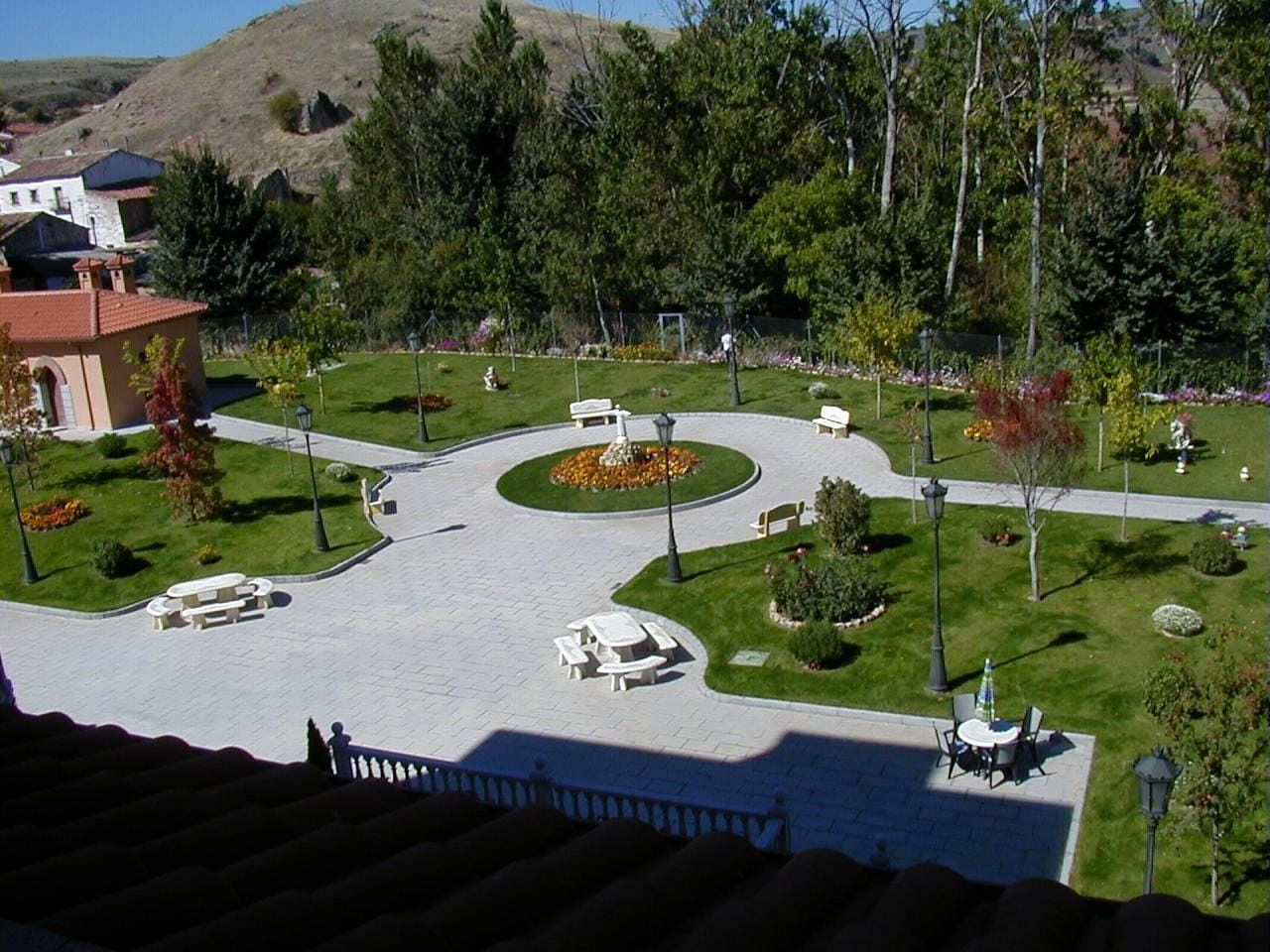 Un parque bien cuidado con un jardín circular central, senderos, bancos y farolas. Al fondo, árboles y una ladera.