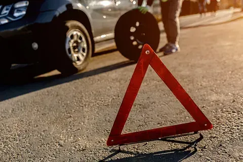Triángulo rojo de emergencia en carretera asfaltada, cambio de rueda pinchada en curso, persona en el fondo, iluminado por el sol.