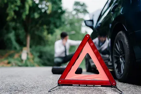 Un triángulo rojo de advertencia en la carretera. Un hombre cambia una llanta; otro habla por teléfono.