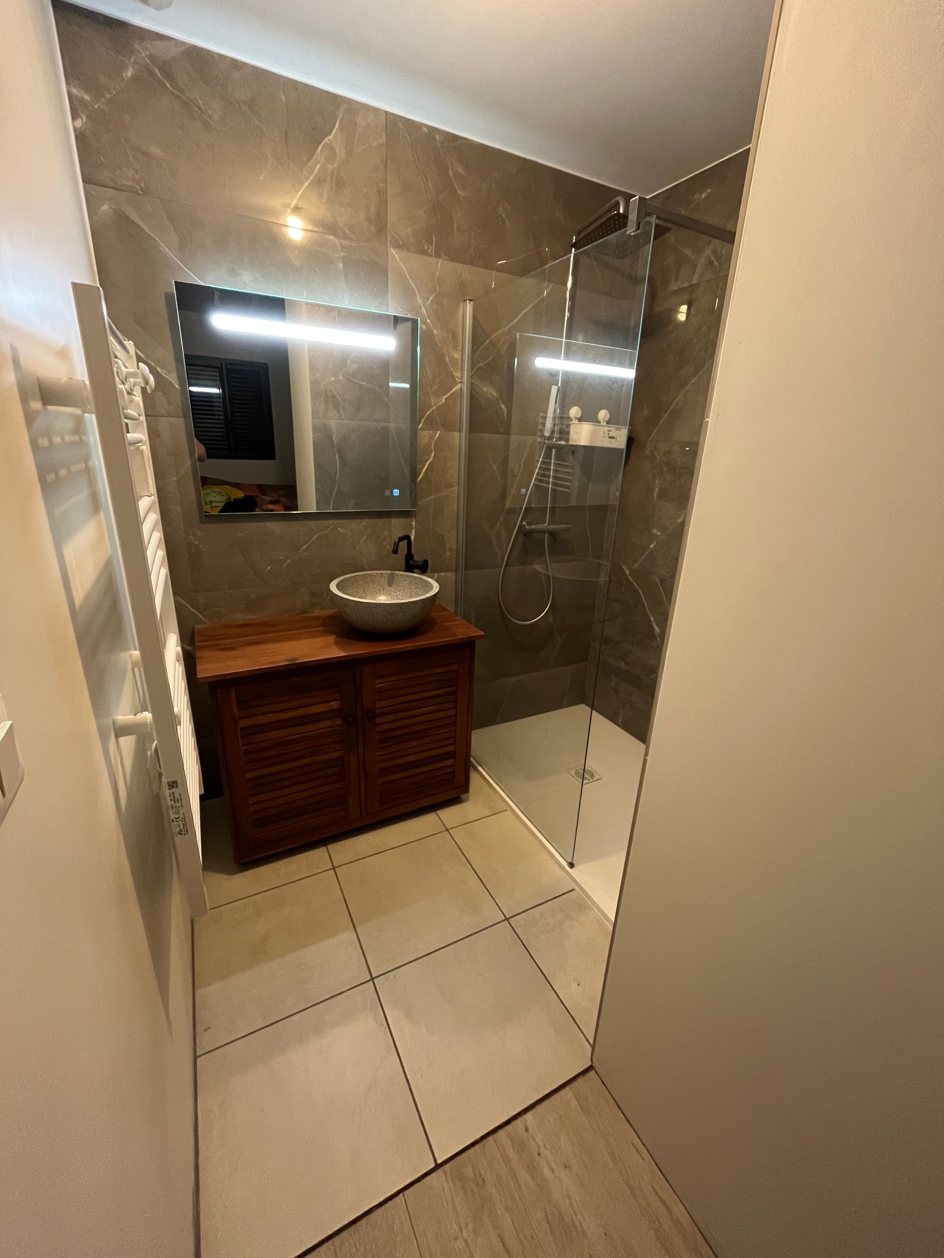 Cabine de douche avec paroi vitrée et carrelage sombre, une vasque sur un meuble en bois à côté