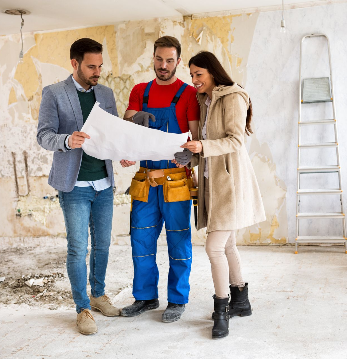 Trois personnes se tiennent dans une pièce en rénovation, regardant un plan tenu par un entrepreneur en salopette bleue.