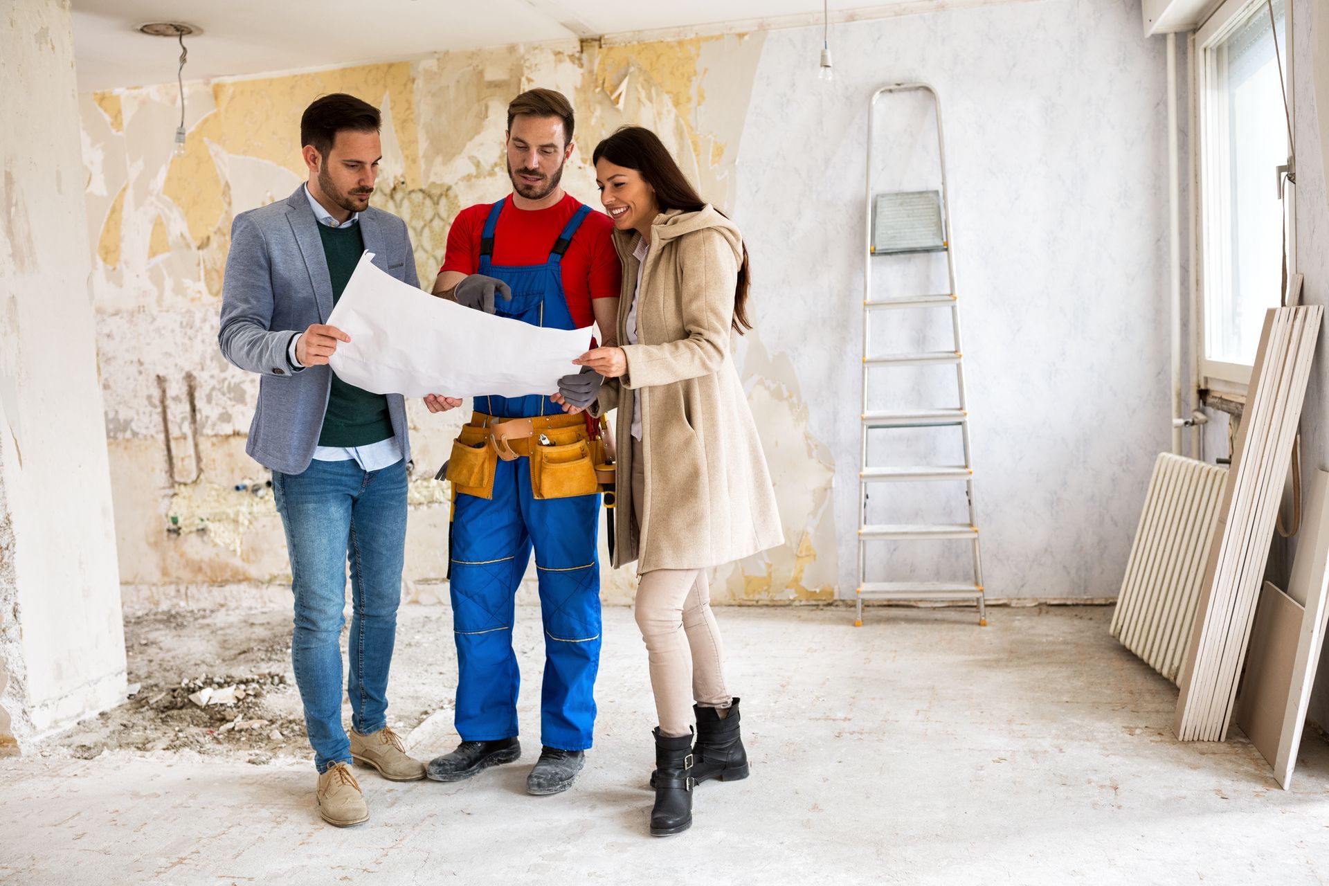 Dans une pièce, une personne en blazer professionnel, un ouvrier en salopette bleue et une femme examinent des plans de construction.