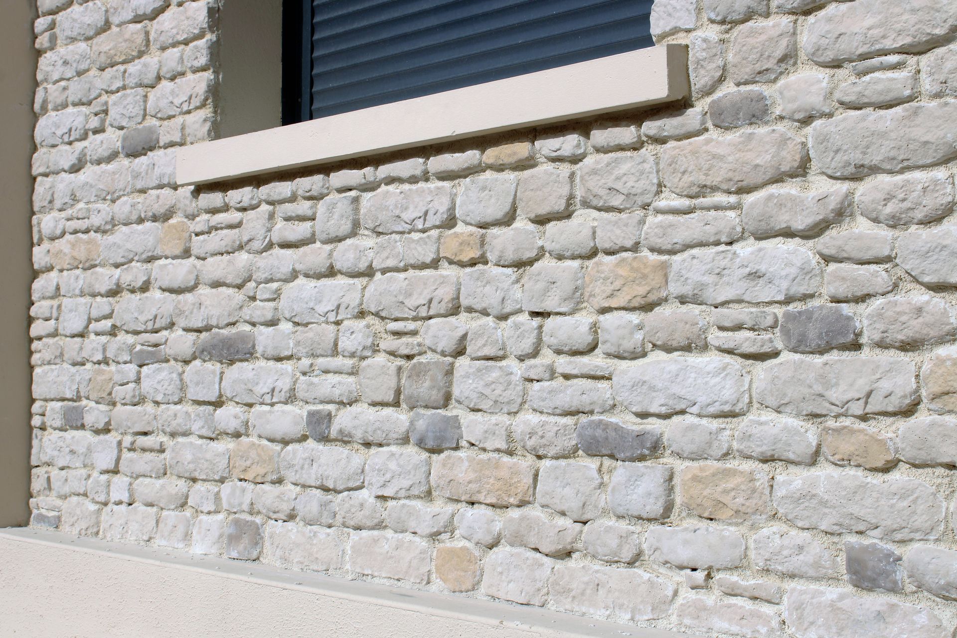 Une façade en pierre claire présente des pierres brutes, irrégulières, de couleur beige et grise, sous une fenêtre à appui beige.