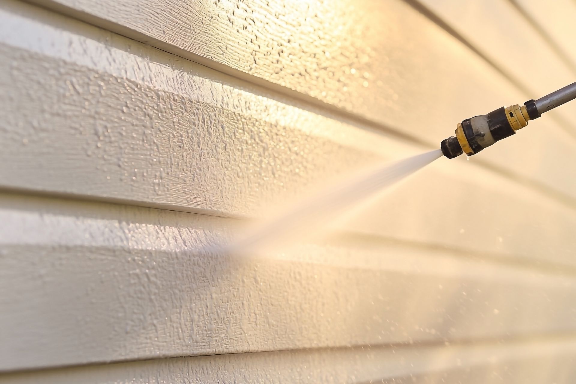Une buse de nettoyeur à haute pression projette de l'eau sur le bardage horizontal beige d'une maison pour en nettoyer la surface.
