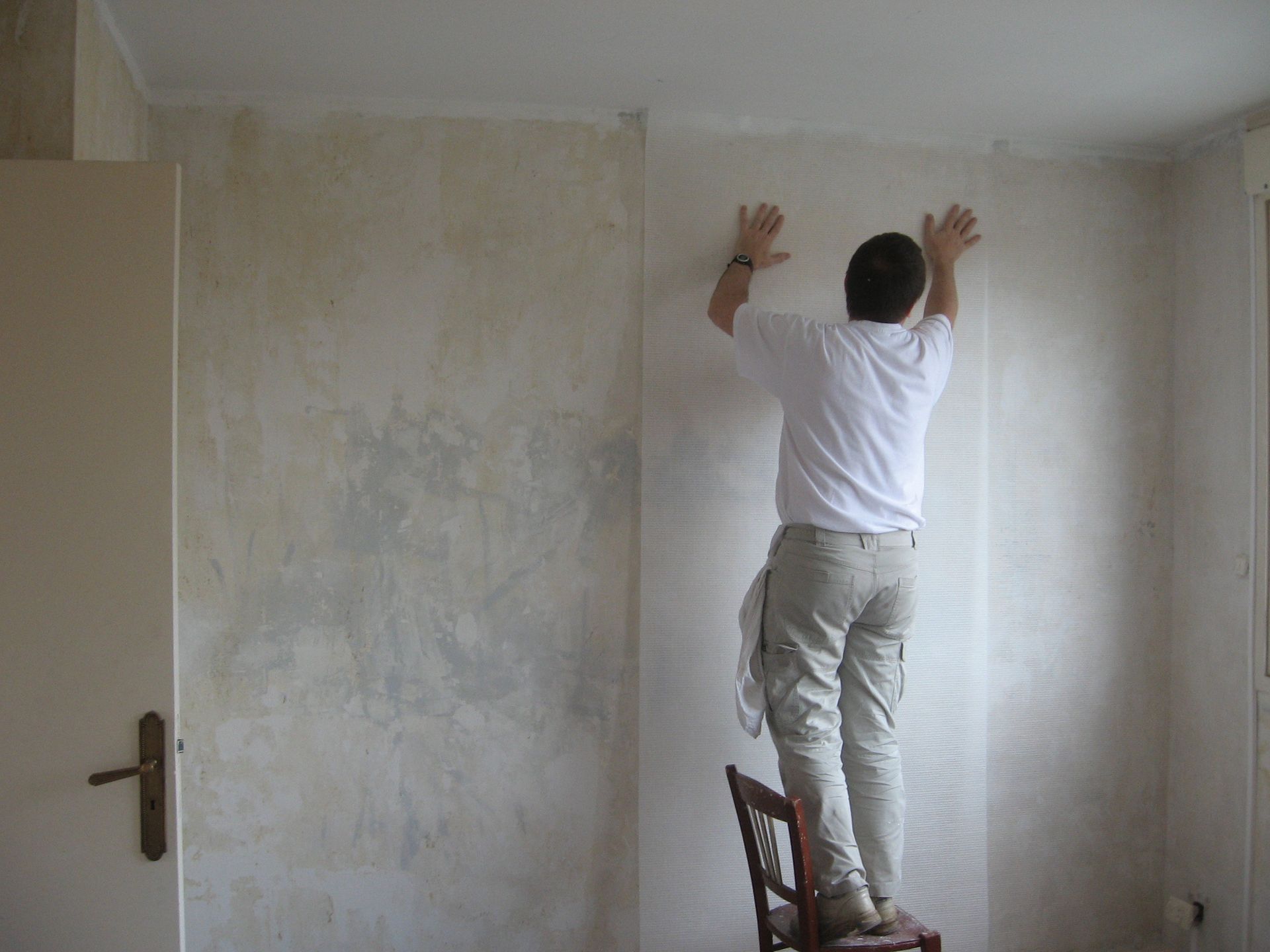 Un homme debout sur une chaise en bois, appliquant un lé de papier peint sur le mur d'une pièce.