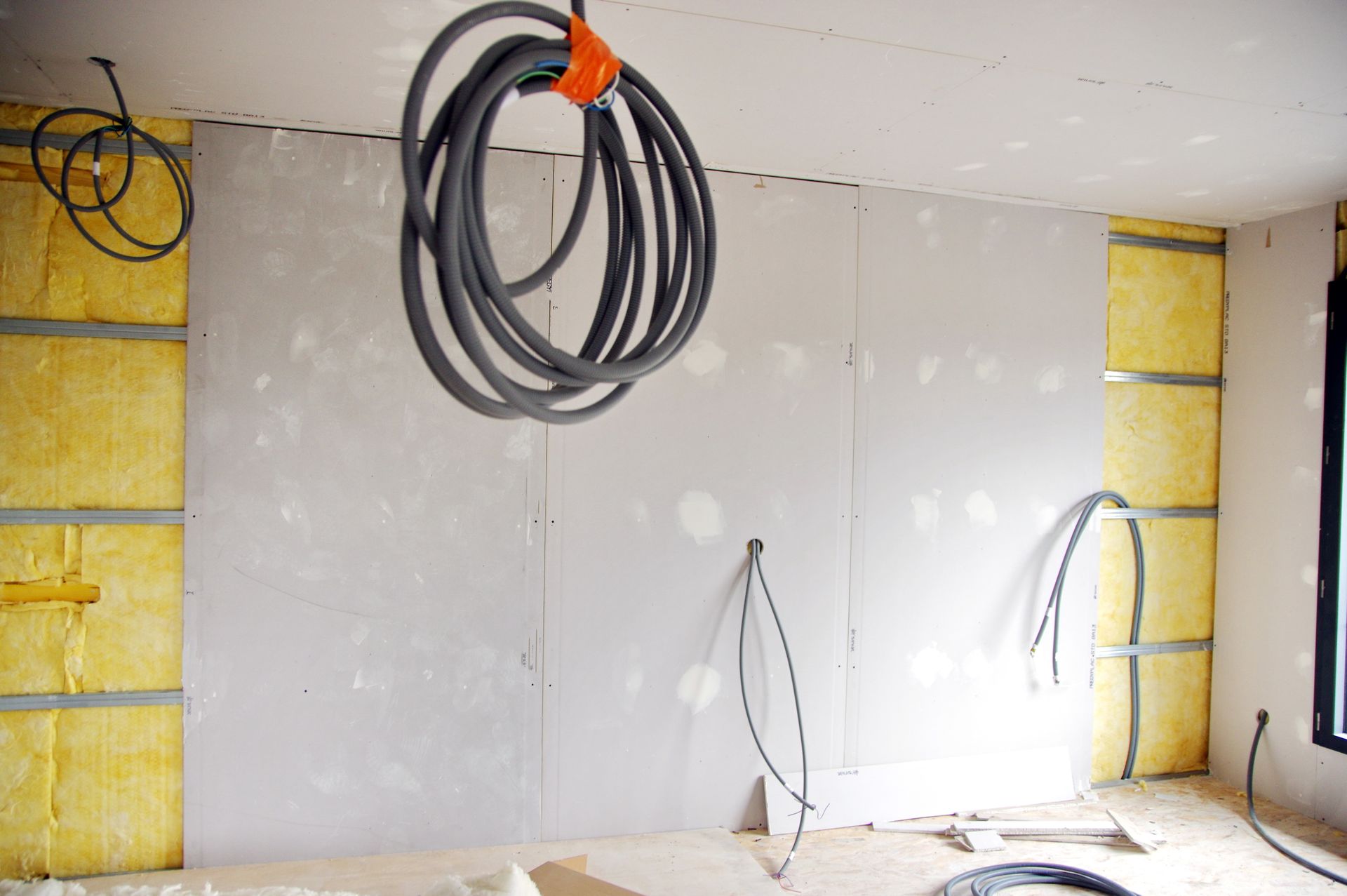 Une pièce intérieure en construction avec des panneaux de plaques de plâtre, de l'isolant apparent et des fils électriques pendants.