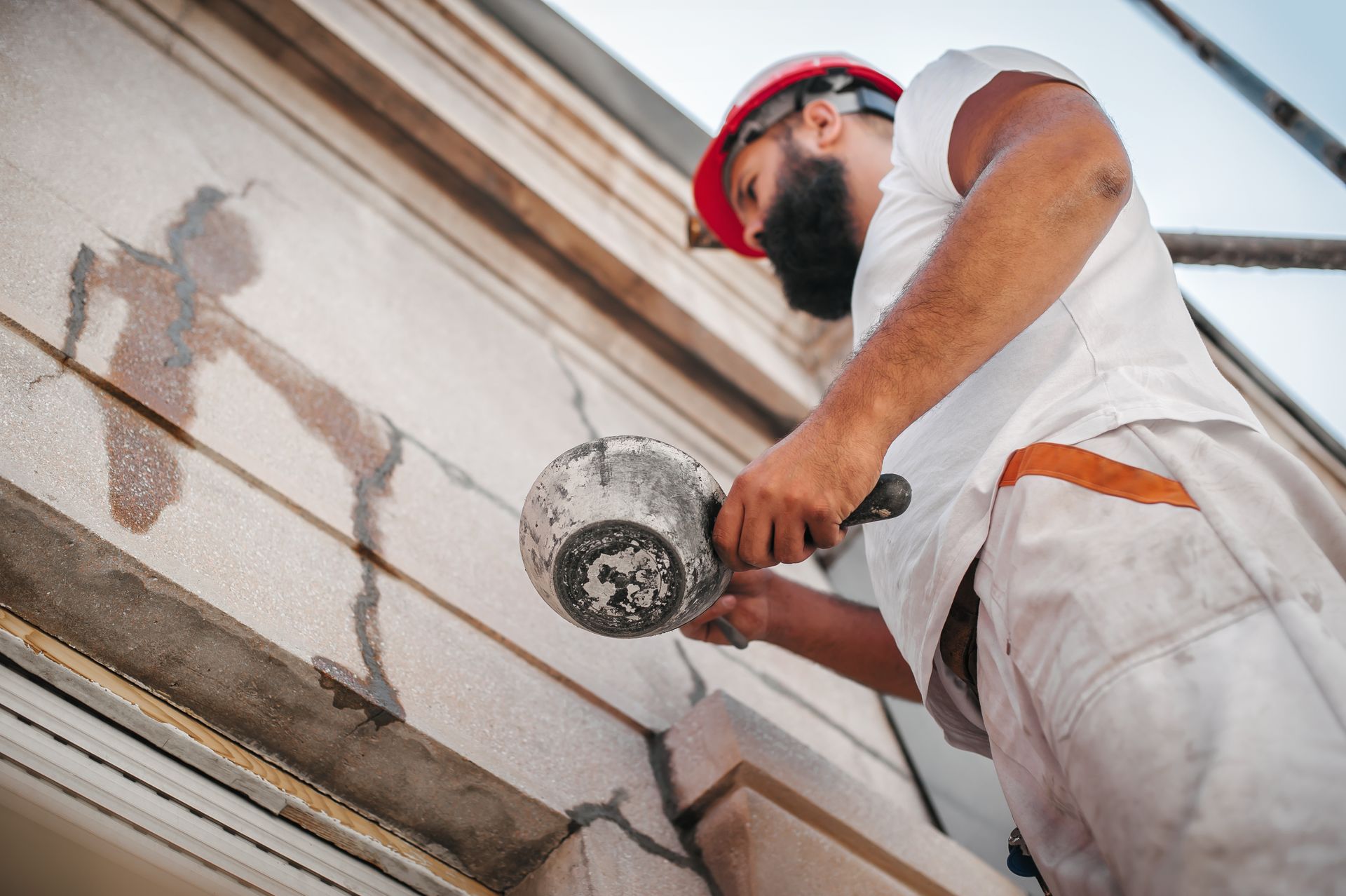 Un ouvrier du bâtiment, portant un casque de chantier, applique du mortier sur la façade en pierre fissurée d'un immeuble.
