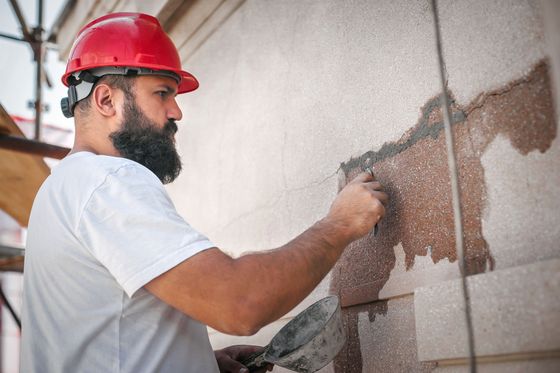 Un ouvrier portant un casque rouge utilise un outil pour appliquer du plâtre ou du mortier sur une partie de la façade d'un bâtiment abîmé par les intempéries.