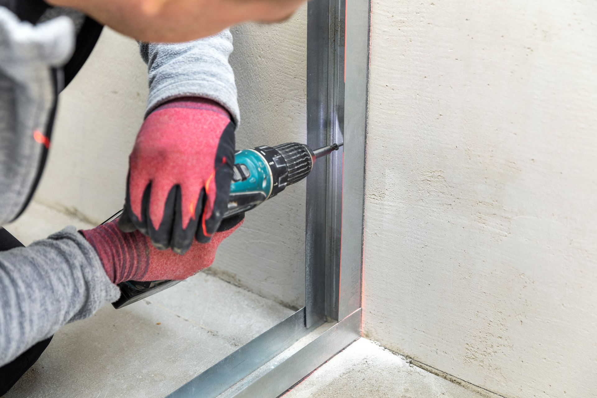 Une personne portant des gants de travail utilise une perceuse électrique pour fixer un montant métallique à un mur.