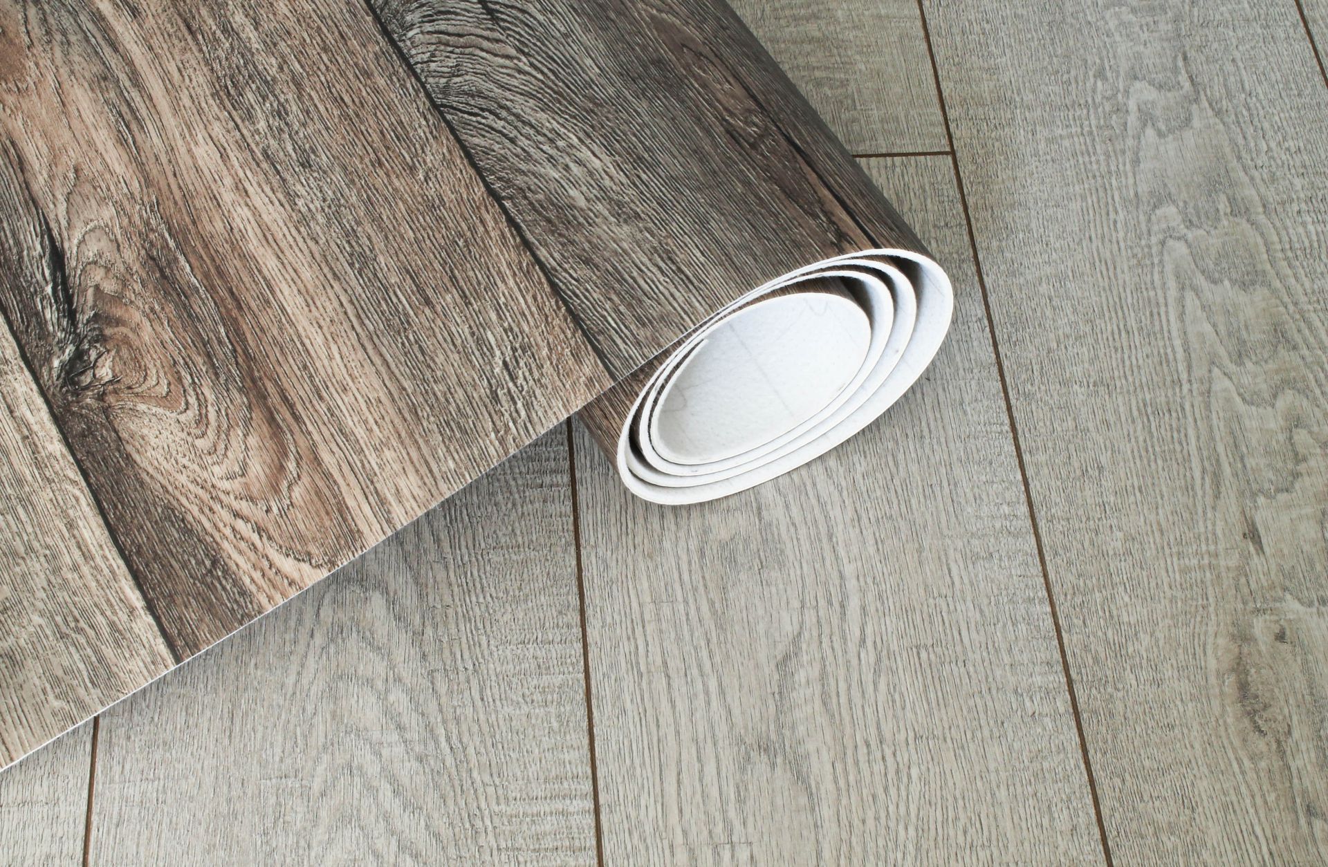 Un rouleau de revêtement de sol en vinyle foncé à motif de grain de bois, posé sur un sol clair gris imitation bois.