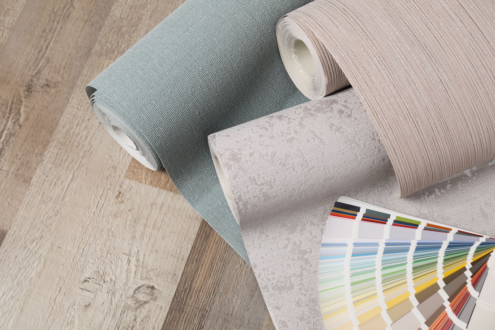 Trois rouleaux de papier peint bleu, beige et gris clair sont posés sur une surface en bois à côté d'un éventail d'échantillons de couleurs.