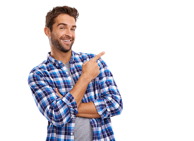 Une personne barbue, vêtue d'une chemise à carreaux bleus par-dessus un T-shirt gris, sourit en pointant du doigt vers la droite.
