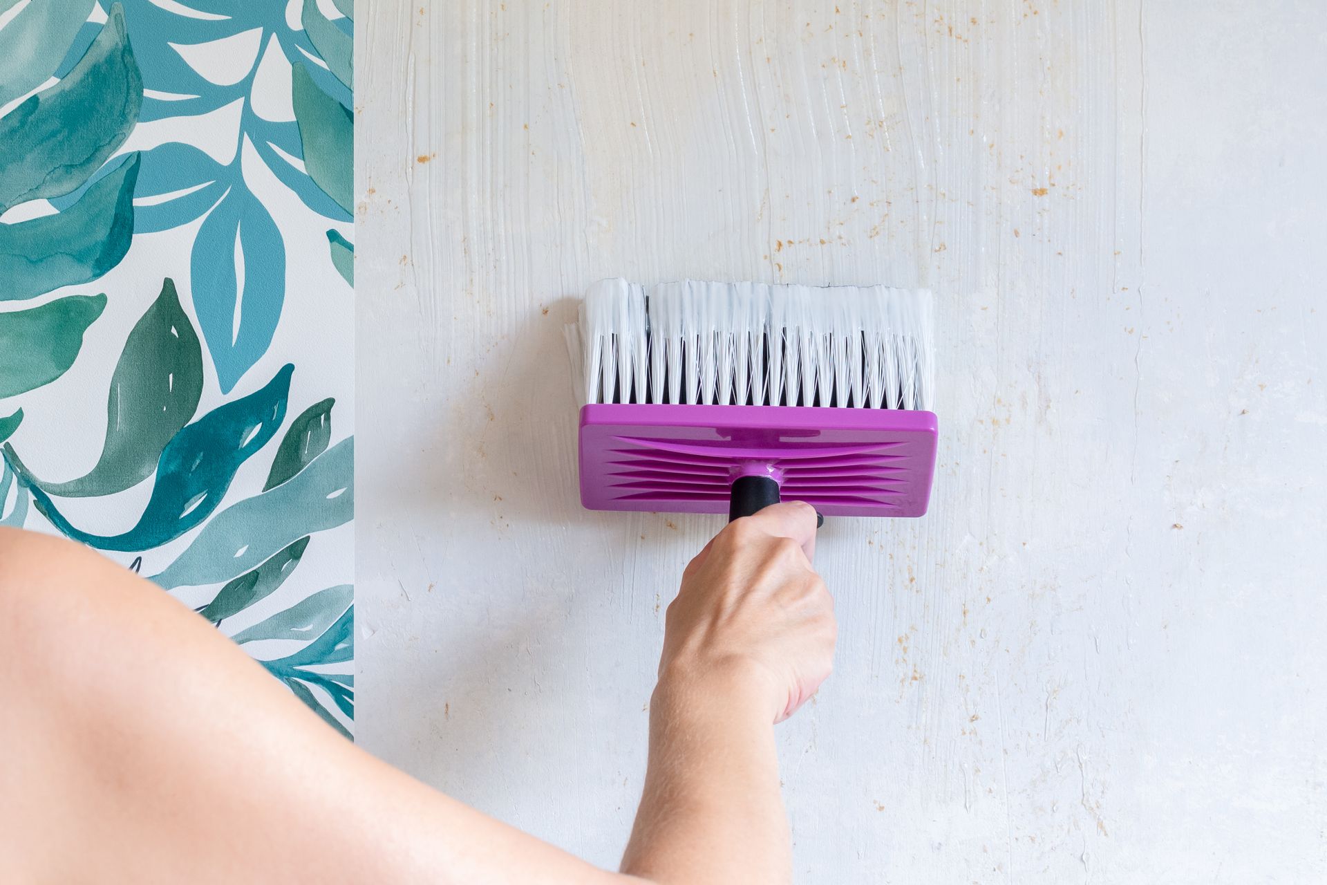 Une main utilise un gros pinceau à manche violet pour appliquer de la colle sur un mur adjacent à un lé de papier peint à motifs botaniques.