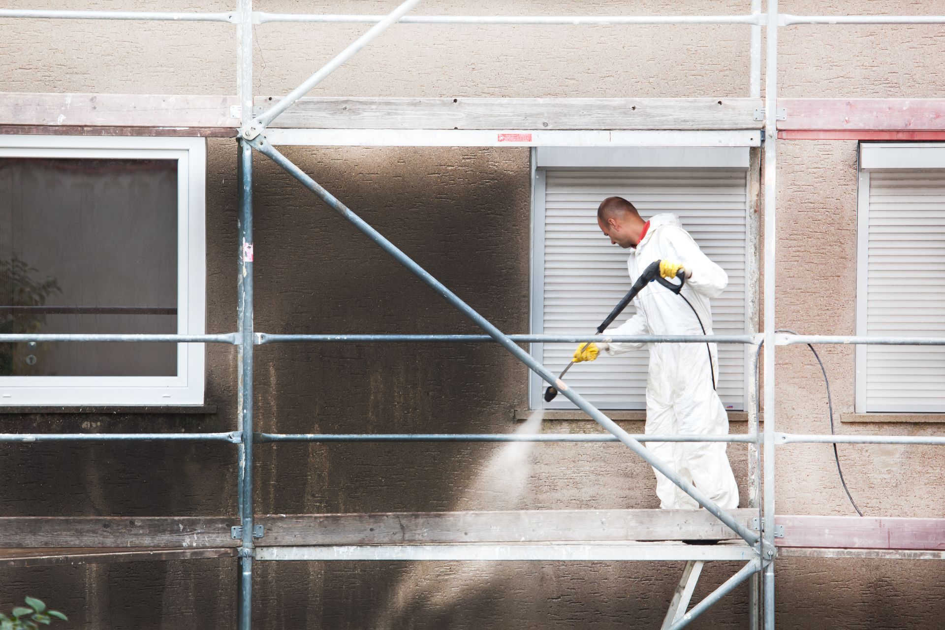 Un ouvrier du bâtiment vêtu d'une combinaison de protection blanche nettoie au jet d'eau à haute pression le mur sombre et taché d'un bâtiment depuis une plateforme d'échafaudage métallique.