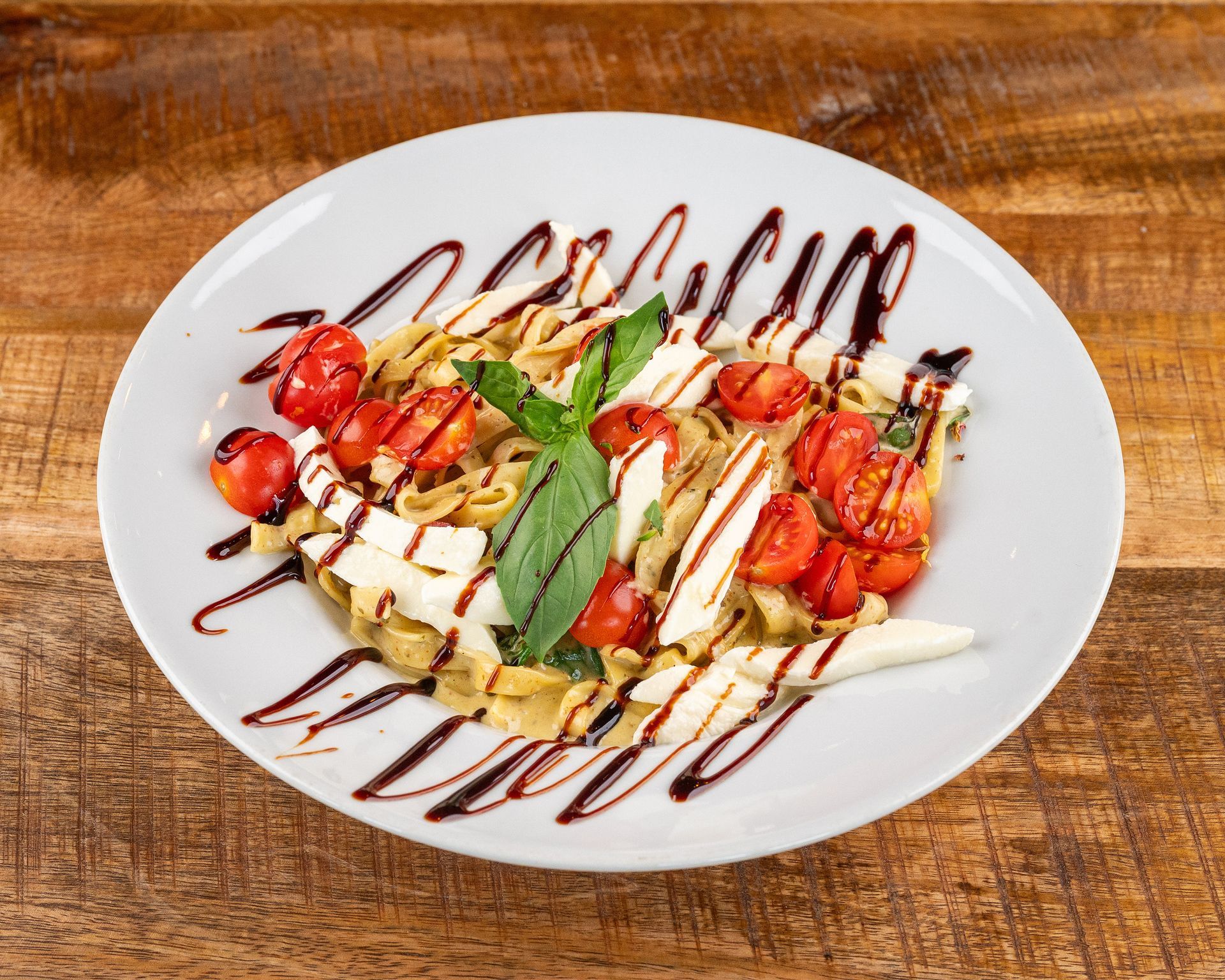Pastagericht mit Tomaten, Mozzarella, Basilikum und Balsamico-Glasur auf einem weißen Teller.