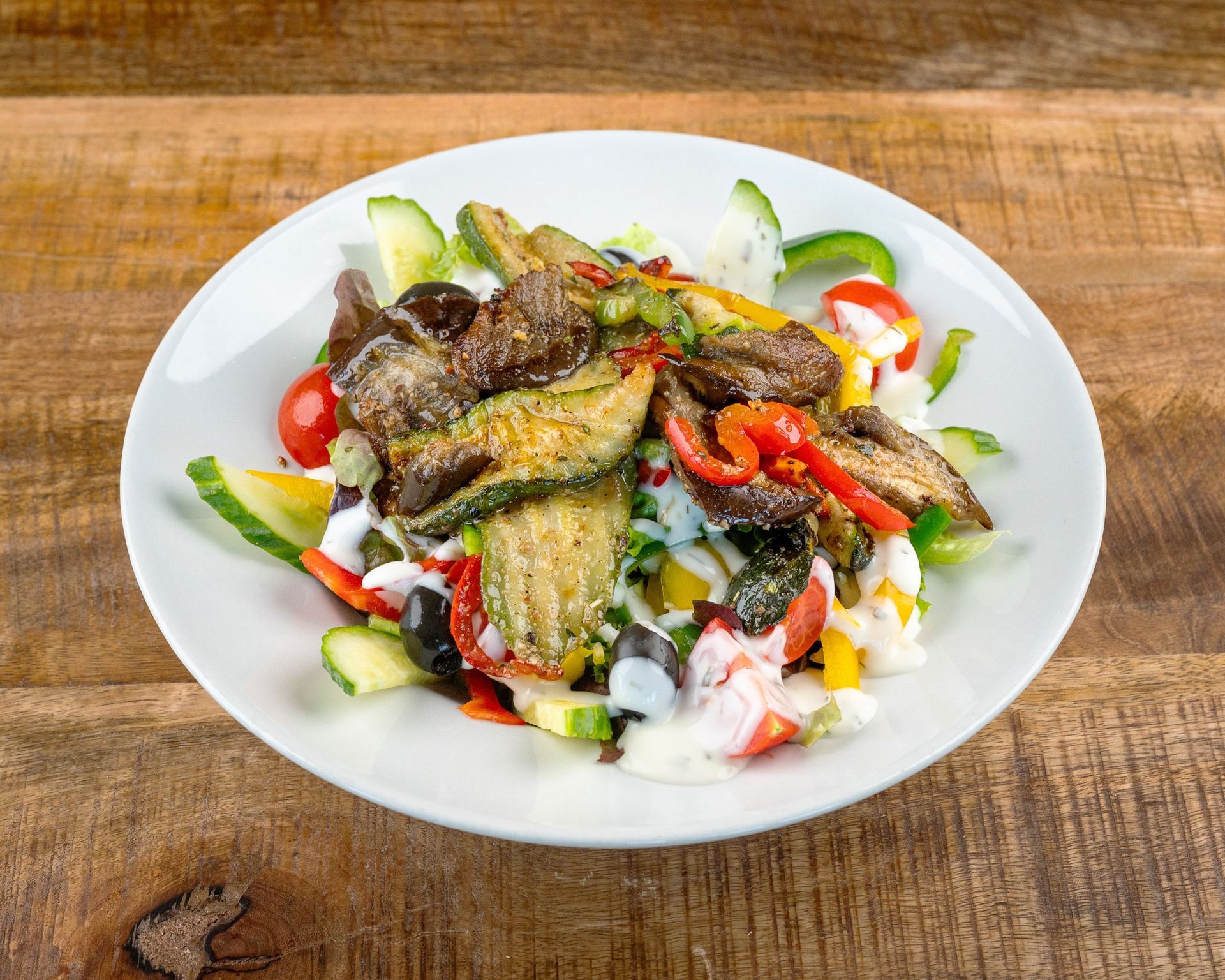 Salat mit gegrillten Zucchini, Tomaten, Oliven, Paprika und Dressing auf einem weißen Teller auf Holz.