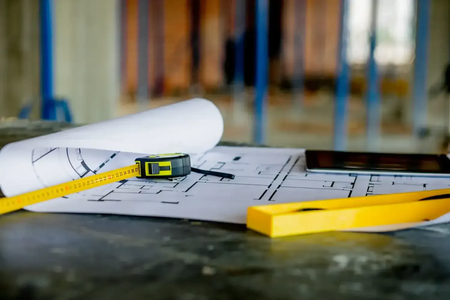 Una cinta métrica amarilla y un nivel apoyados sobre planos arquitectónicos en una obra en construcción.
