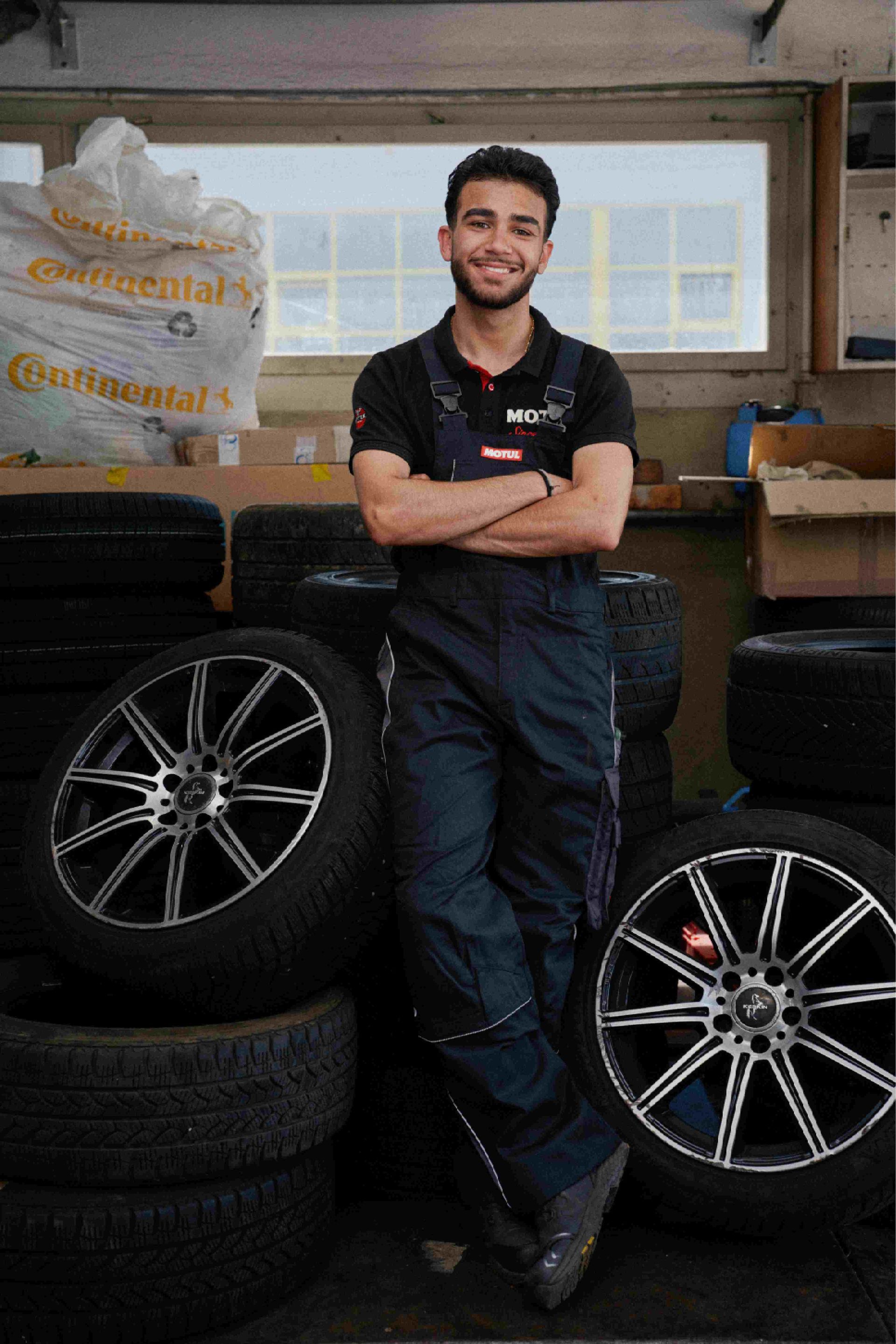 Mann in Arbeitsuniform, lächelnd, mit verschränkten Armen, lehnt in einer Garage an Reifen.
