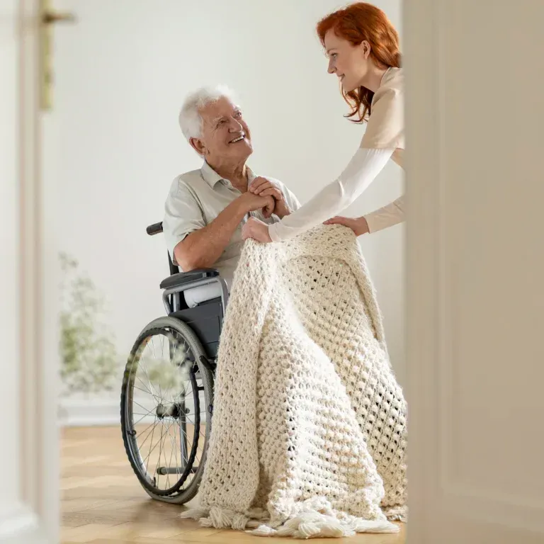 Una mujer sostiene una manta sobre un anciano en silla de ruedas.
