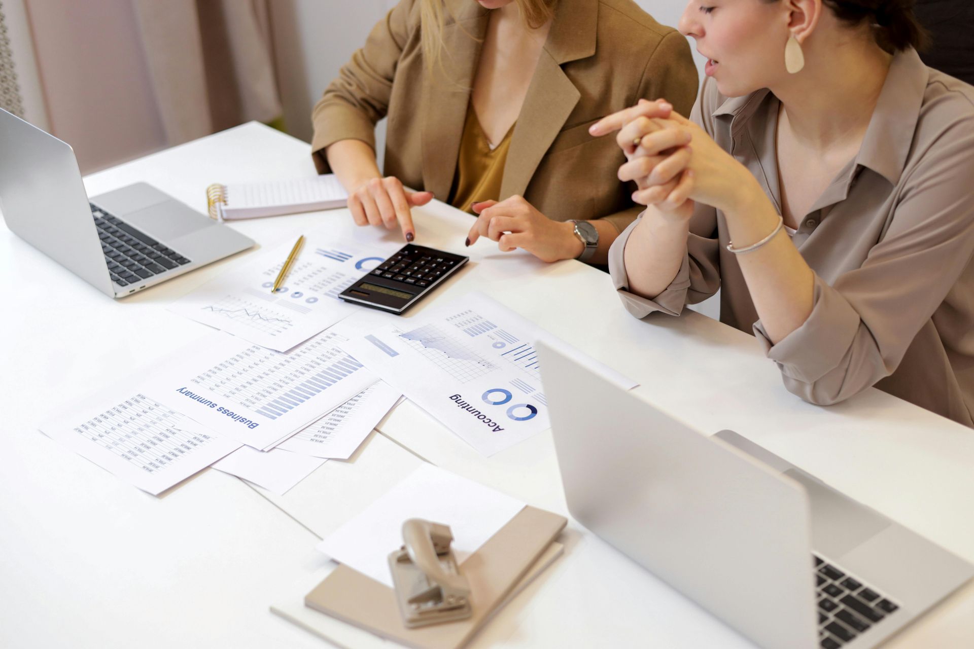 Duas mulheres sentadas à mesa com laptops, documentos e uma calculadora, discutindo dados financeiros.
