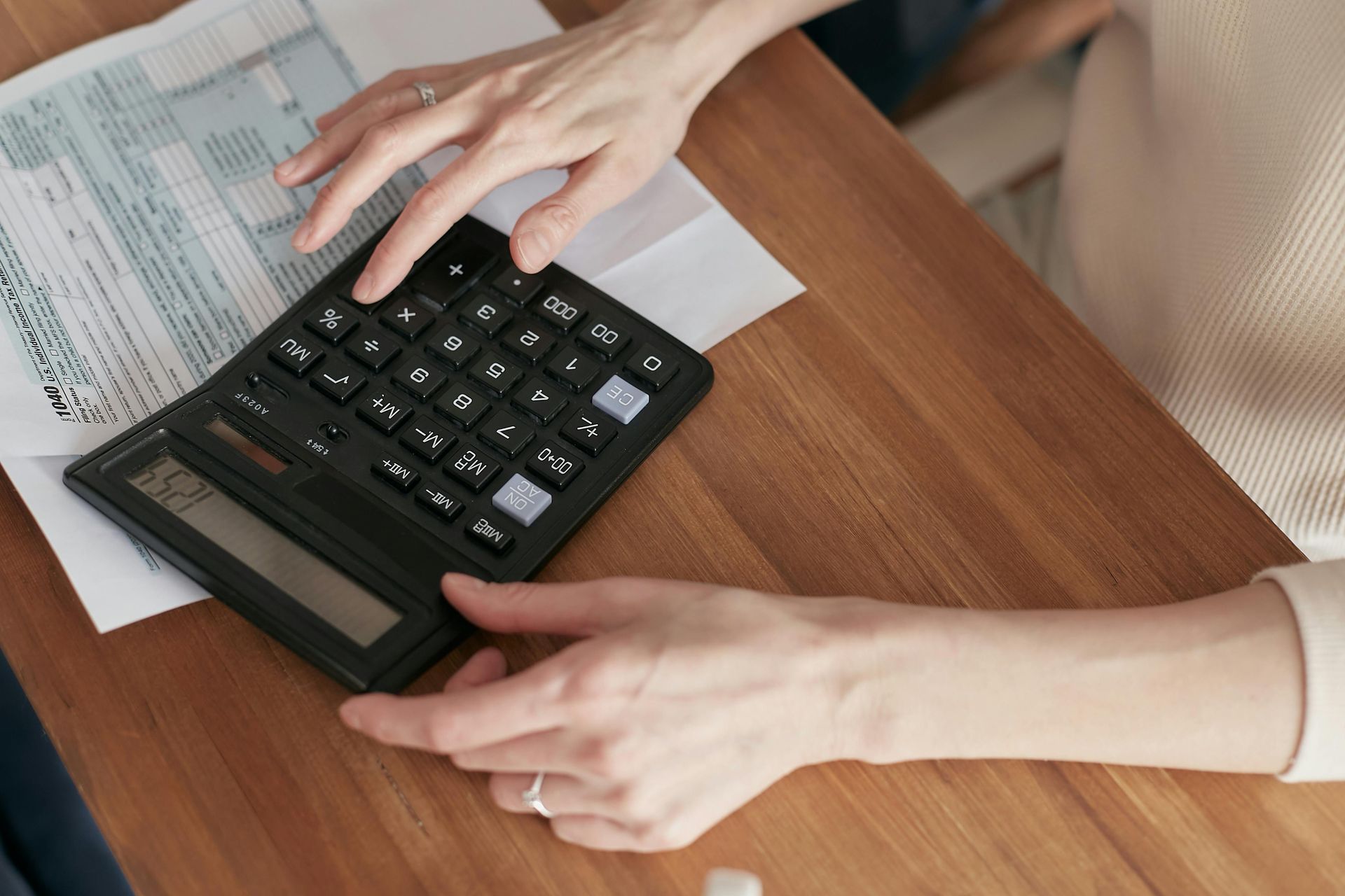 Mãos de uma pessoa usando uma calculadora preta sobre uma mesa de madeira com papéis por perto.