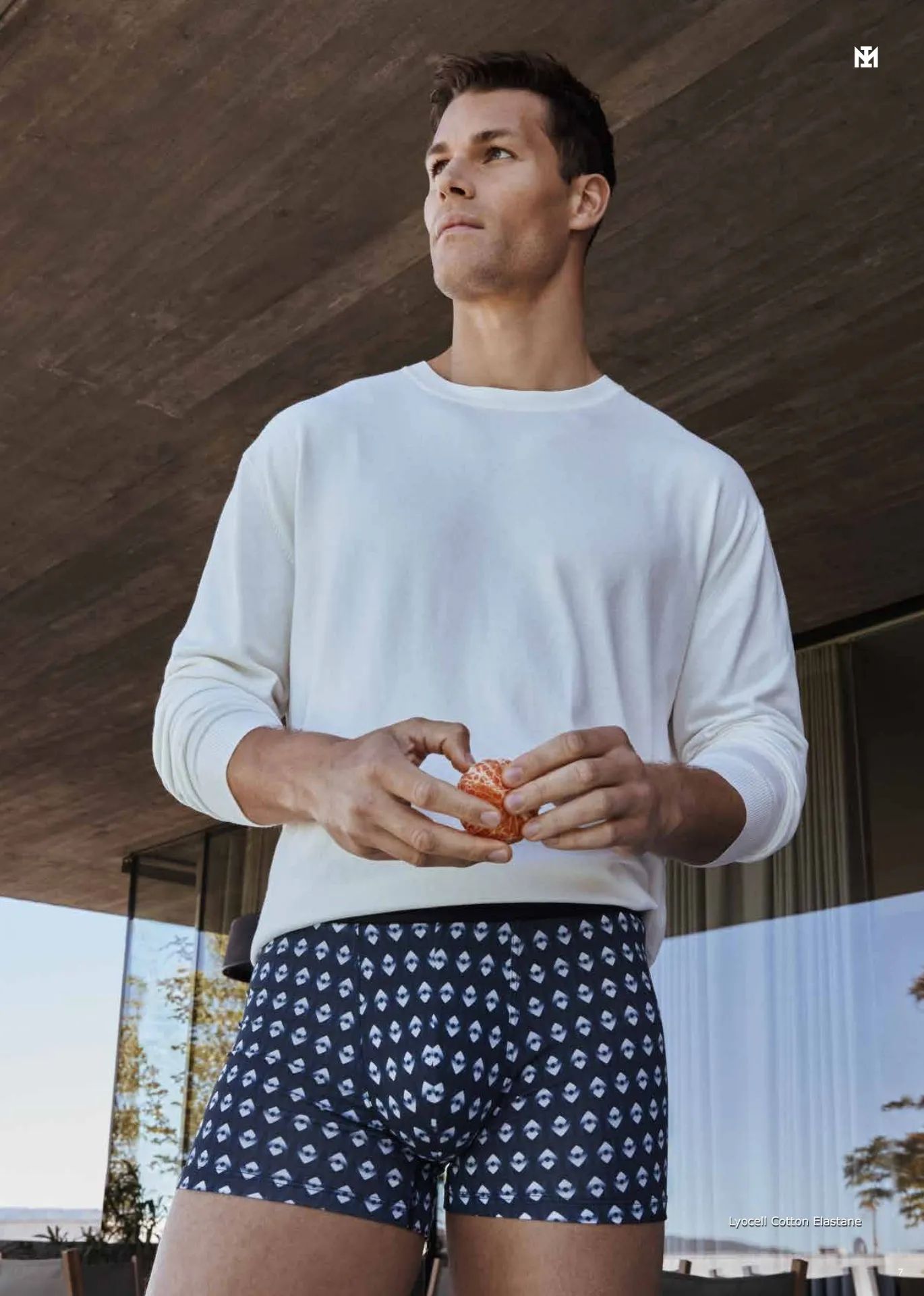 Un hombre que viste un suéter blanco y calzoncillos azules está de pie en un balcón.