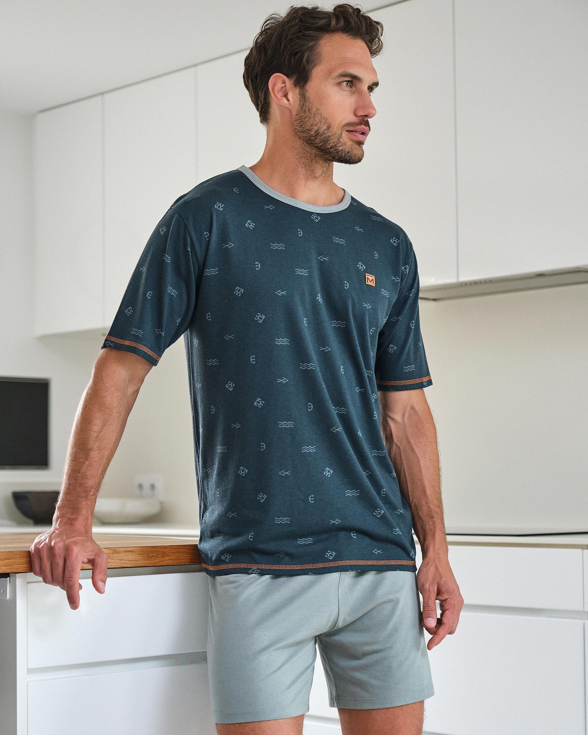 Un hombre con una camisa azul y pantalones cortos grises está de pie en una cocina.