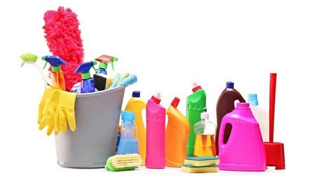 Materiales de limpieza: balde con sprays, guantes, plumeros, botellas y un trapeador, sobre una superficie blanca.