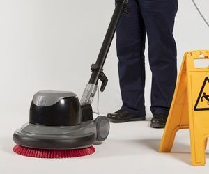 Persona limpiando el piso con una pulidora; hay un cartel amarillo de