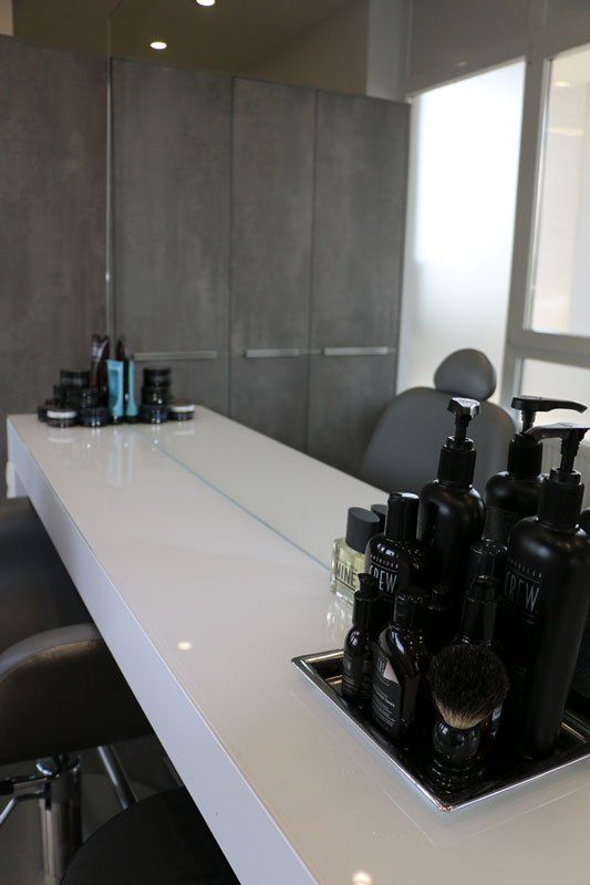 Weißer Tisch in einem Friseursalon mit schwarzen Pflegeprodukten und Spiegel. Graue Wände, Stuhl.