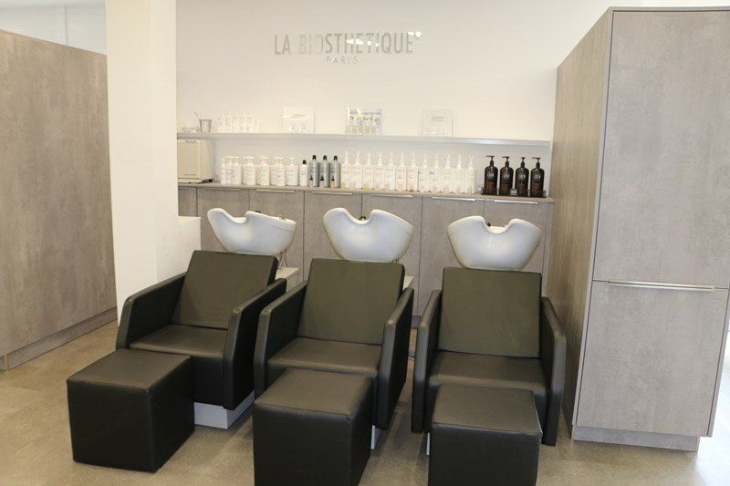 Friseursalon mit drei Waschplätzen. Graue Sessel und Fußstützen stehen vor weißen Waschbecken. Flaschen stehen auf einem Regal.