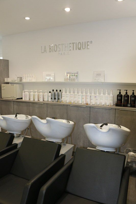 Salon mit Shampoo-Stationen: weiße Becken, schwarze Stühle, Produktregale und der Markenname „La Biosthetique“.