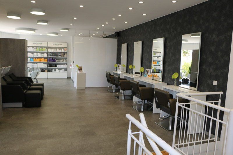 Salon-Innenraum mit Stylistenstationen, Spiegeln und Produktpräsentation vor einer dunkelgrauen strukturierten Wand.