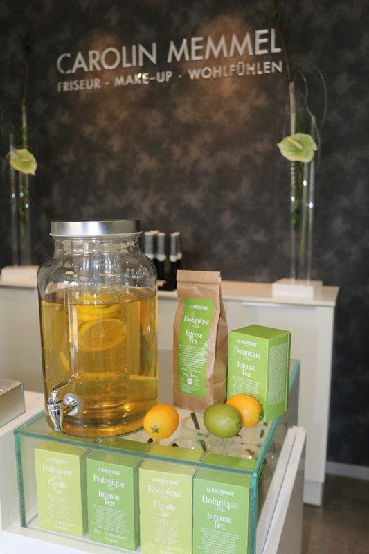 Auslage mit Tee und Zitrusfrüchten in einem Salon, mit einem Getränkespender aus Glas und einem Schild mit der Aufschrift „CAROLIN MEMMEL“