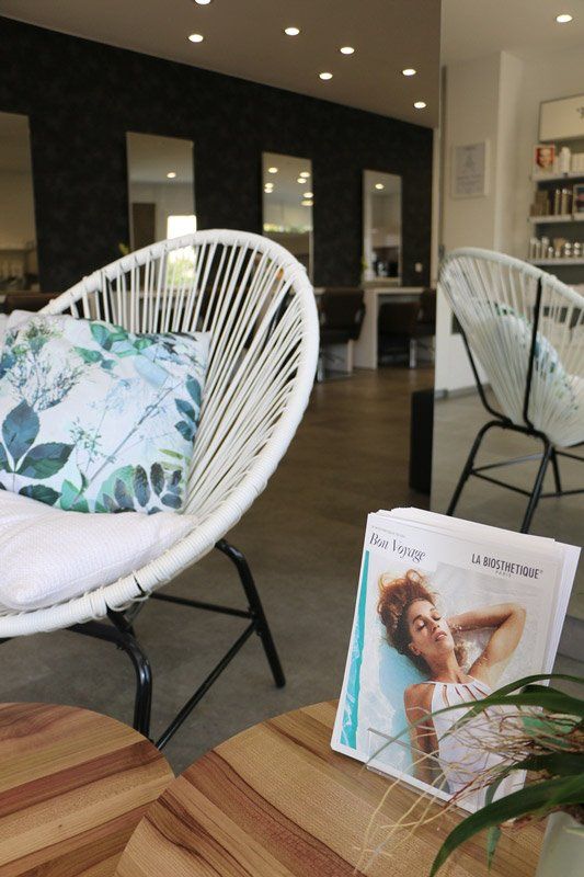 Ein Wartebereich in einem Friseursalon mit weißen geflochtenen Stühlen, einem Kissen und Zeitschriften auf einem Holztisch.