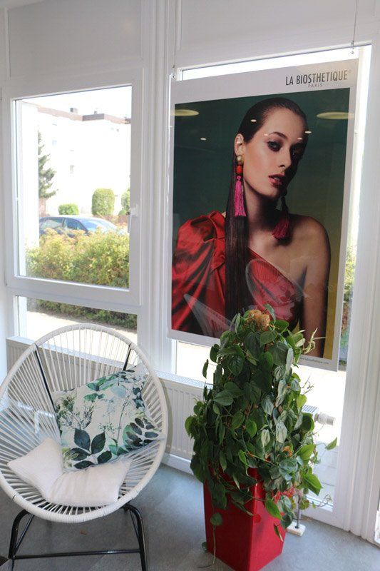 Ein Modeposter für Damen neben einem Fenster mit einem weißen Stuhl und einer großen Pflanze.