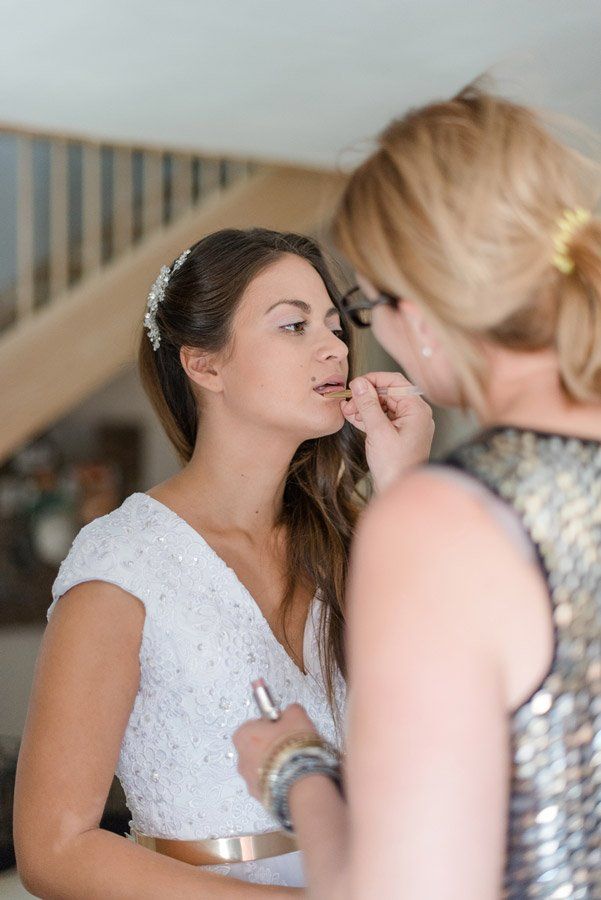 Braut lässt sich von einem Maskenbildner Lippenstift auftragen; beide sind im Innenbereich, in der Nähe einer Treppe.