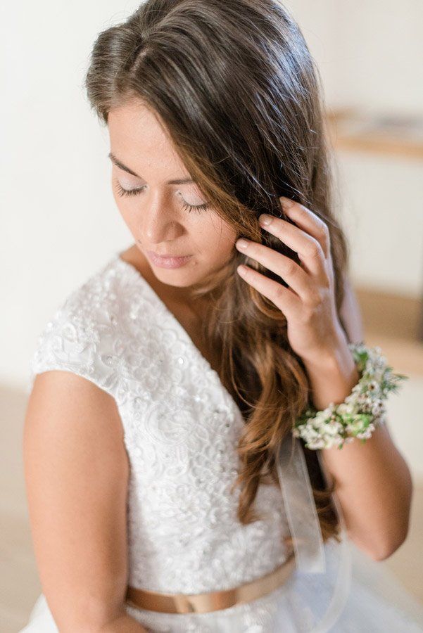 Braut im weißen Spitzenkleid, berührt ihr Haar und blickt nach unten. Grün-weißes Blumenarmband am Handgelenk.