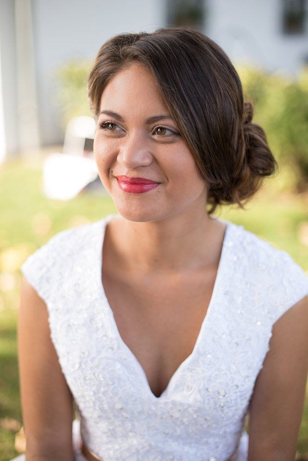 Frau im weißen Kleid mit Hochsteckfrisur, rotem Lippenstift, lächelnd im Freien.
