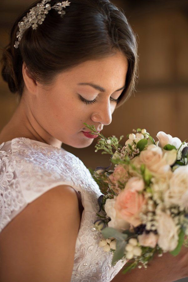 Braut im weißen Spitzenkleid, die einen Rosenstrauß duftet, drinnen mit einem dekorativen Kopfschmuck.