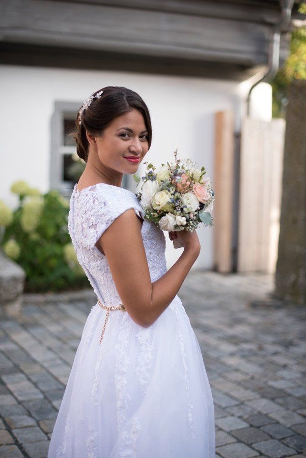 Braut hält einen Blumenstrauß, lächelt und blickt über ihre Schulter vor einem weißen Gebäude.
