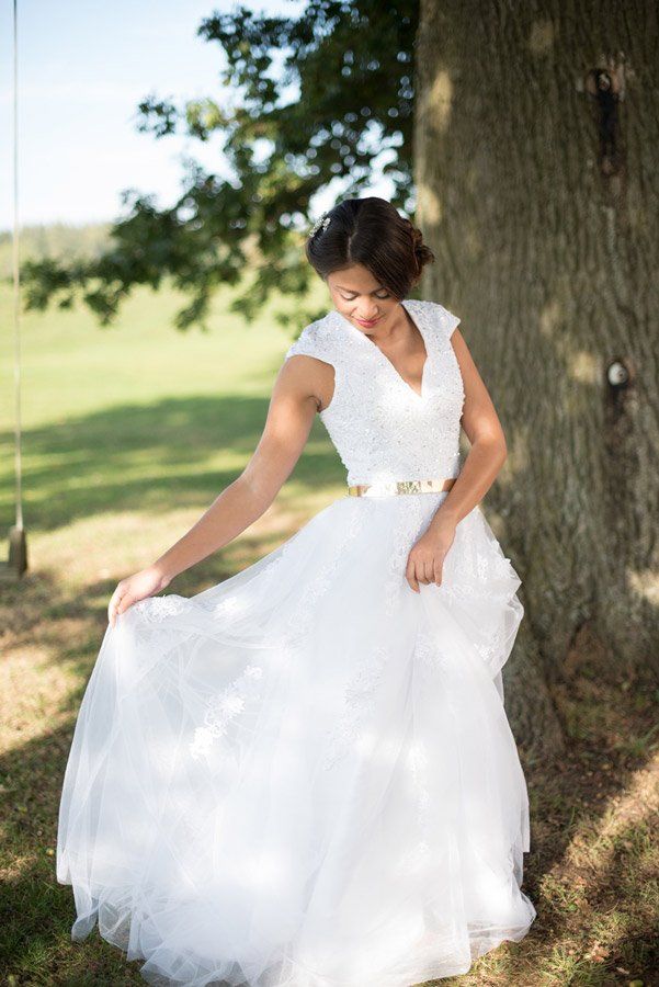 Braut im weißen Kleid mit goldenem Gürtel posiert neben einem Baum auf einem Feld.