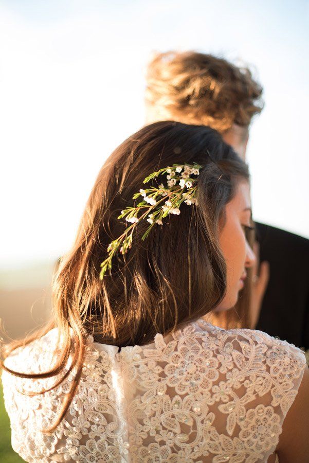 Braut mit floralem Haarteil; Bräutigam im Hintergrund. Frau trägt ein Spitzenkleid und blickt nach unten.