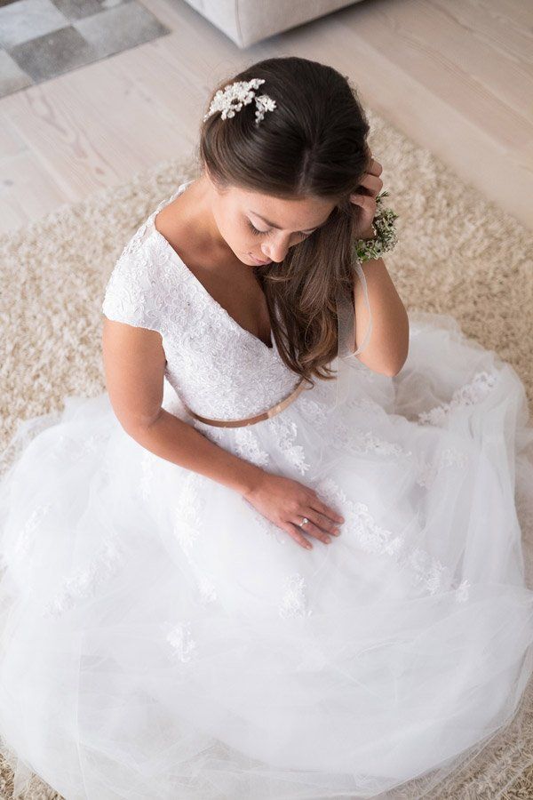 Braut im weißen Kleid mit goldenem Gürtel sitzt auf cremefarbenem Teppich und blickt nach unten.